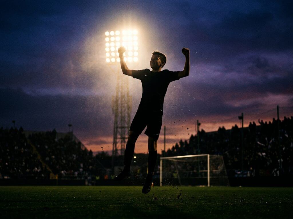 Youth soccer player celebrating goal under stadium lights at sunset.